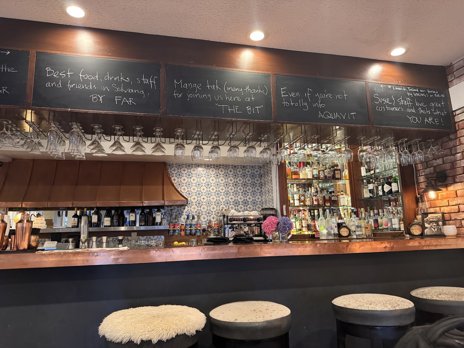 Warm bar interior with copper details, hanging glassware, chalkboards, and bottle displays